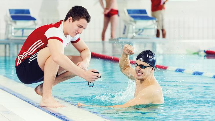 Schwimmtrainer zeigt Stoppuhr, Sportler freut sich über die Zeit.